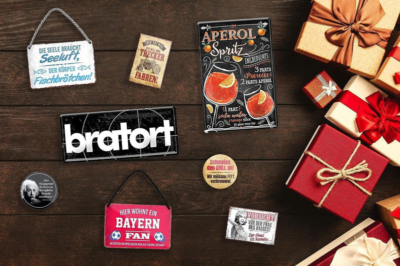 Das Bild zeigt verschiedene Dekostücke und Geschenkideen wie Magnete, Schilder und Etiketten. Darauf zu sehen sind Marken wie "Bratari" und "Aperol Spritz" sowie Sprüche wie "Rinderfiletarier". Diese Elemente sind auf einem dunklen, hölzernen Hintergrund platziert, zusammen mit roten Geschenkverpackungen. Insgesamt vermittelt das Bild eine gemütlich-dekorative Atmosphäre.